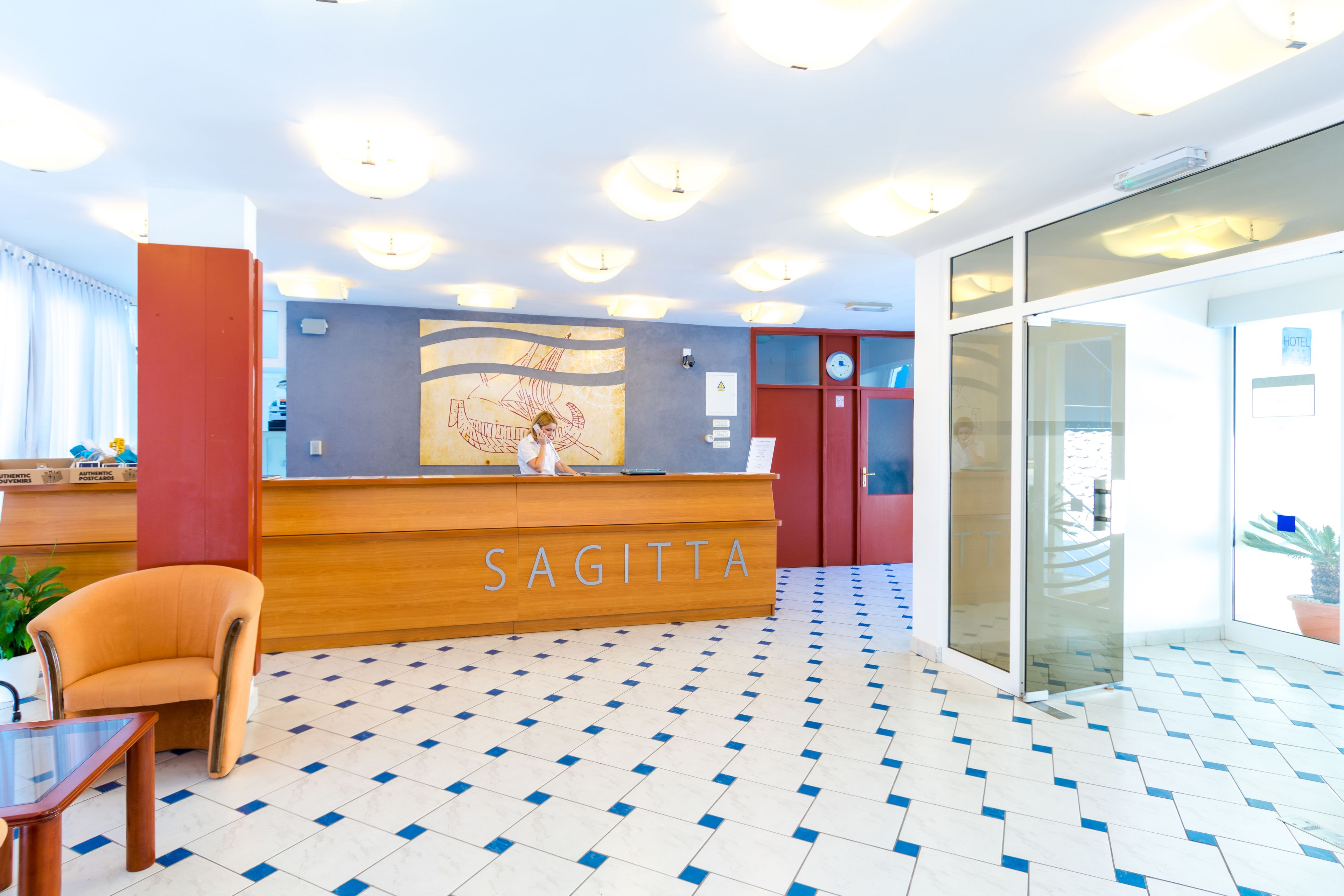 A hotel reception area with a check-in counter labeled "SAGITTA," a modern design with a patterned floor, and a glass door leading outside.