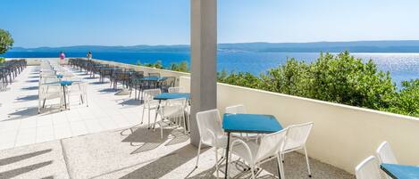 An outdoor terrace with white chairs and tables, overlooking a scenic view of the sea and distant hills.