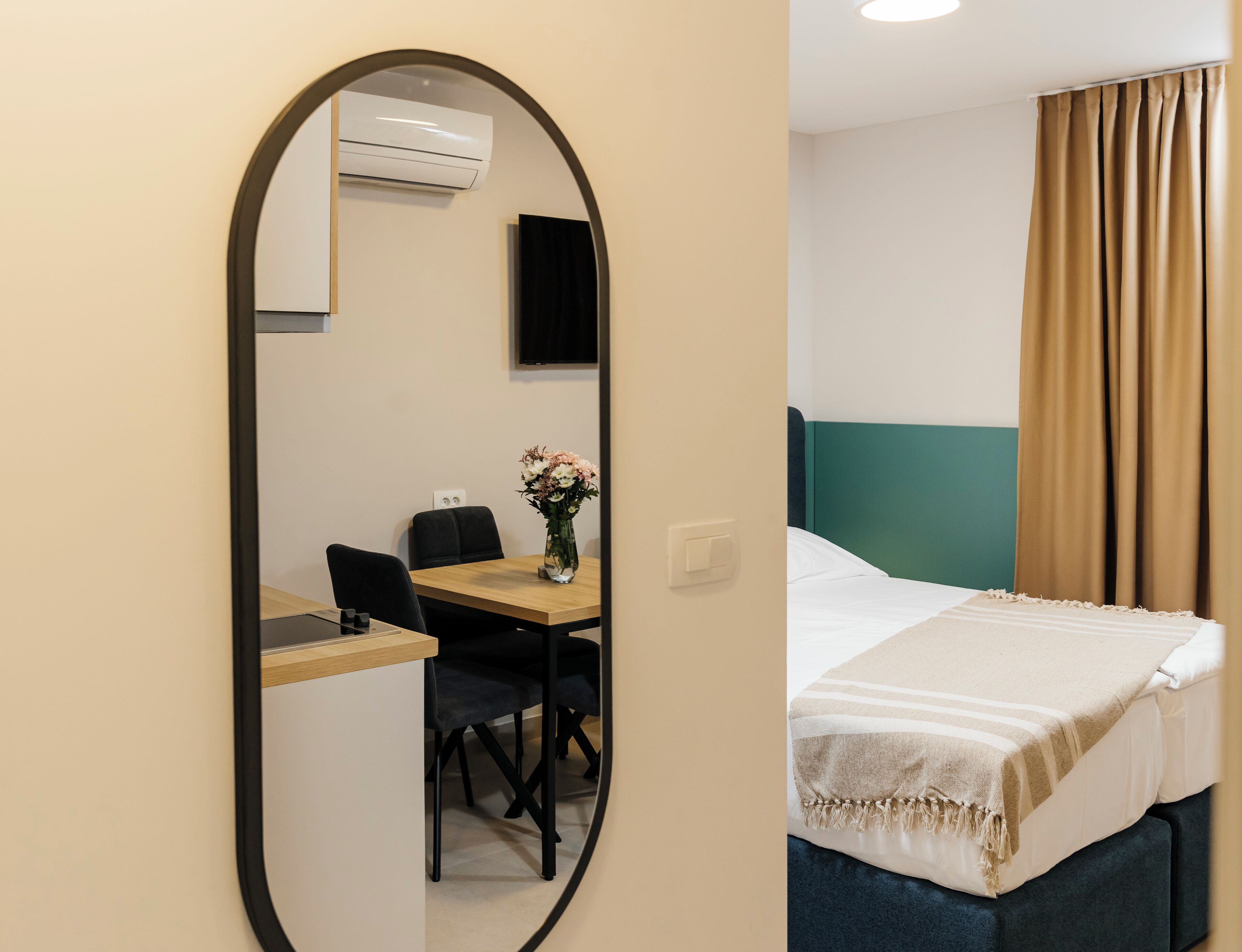 A hotel room with a bed, a desk with a chair, a mirror, and a wall-mounted TV.