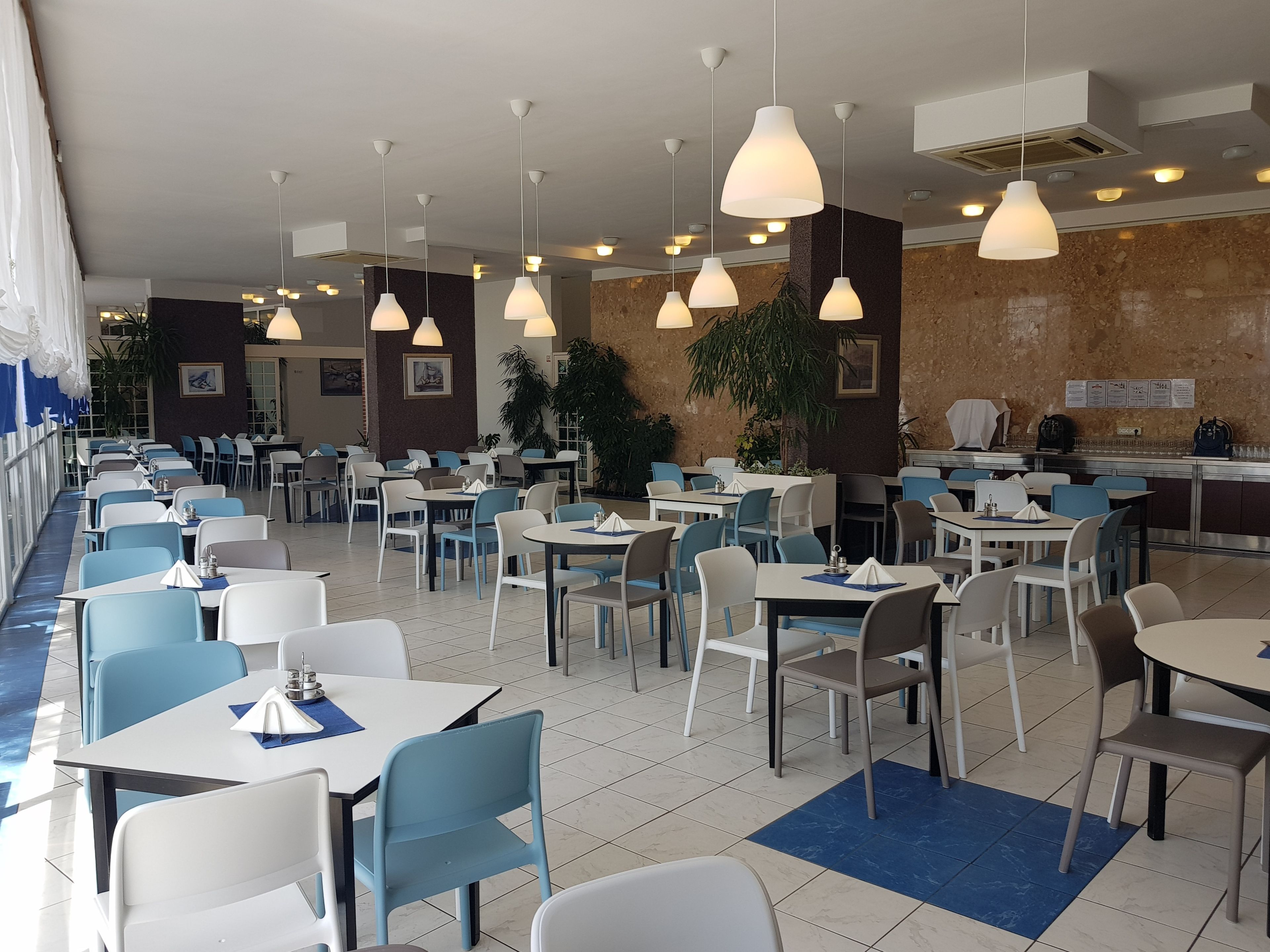 Un restaurante espacioso con numerosas mesas y sillas, lámparas colgantes y zonas de estar tanto en el interior como en el exterior.