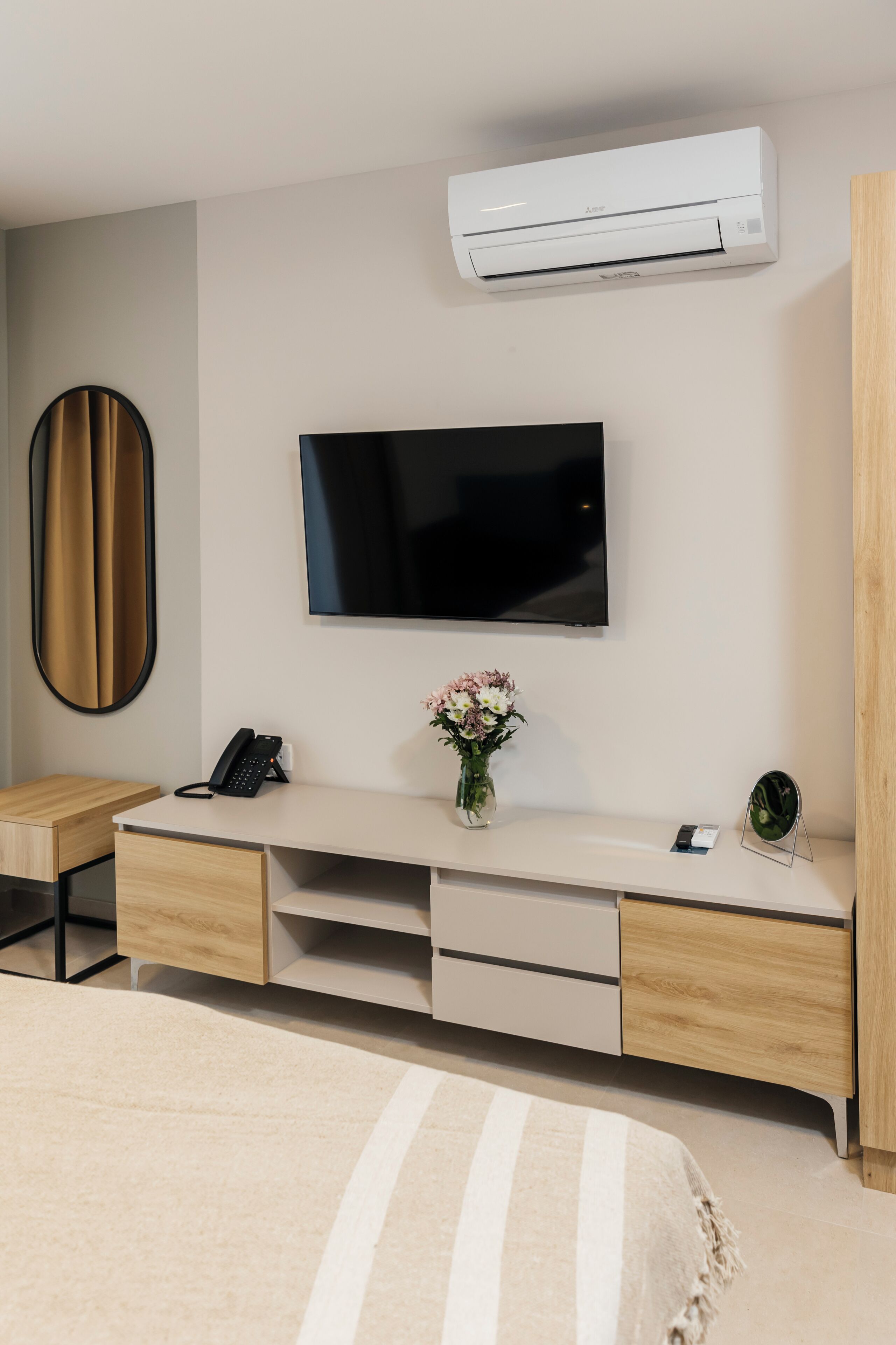 Een moderne hotelkamer met een flatscreen televisie aan de muur, een houten bureau met een telefoon en een bloevas, een bed met een gestreept dekbed en een airconditioner boven de televisie.