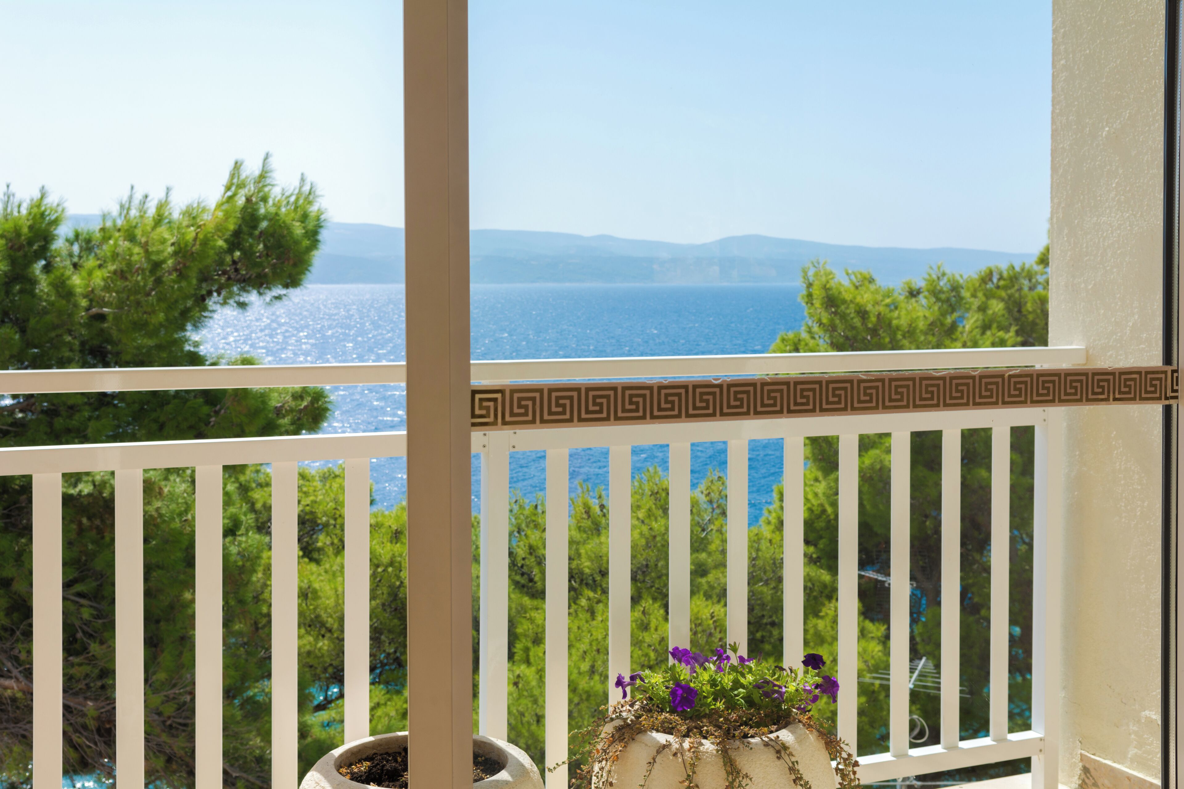 A balcony with a view of the sea and mountains, a potted plant with flowers, and a white railing with a decorative pattern.
