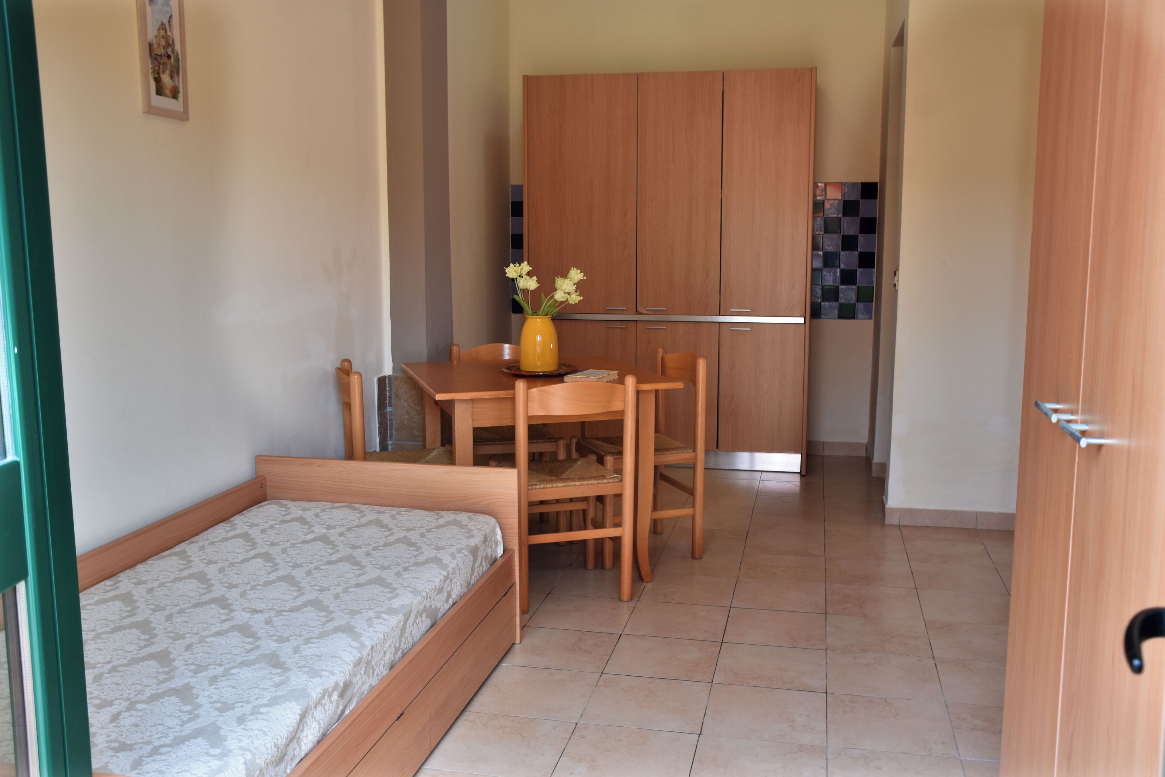Une pièce de taille modeste comprenant un lit simple, une petite table à manger avec des chaises, un coin cuisine avec des placards et une porte dotée d’une poignée.