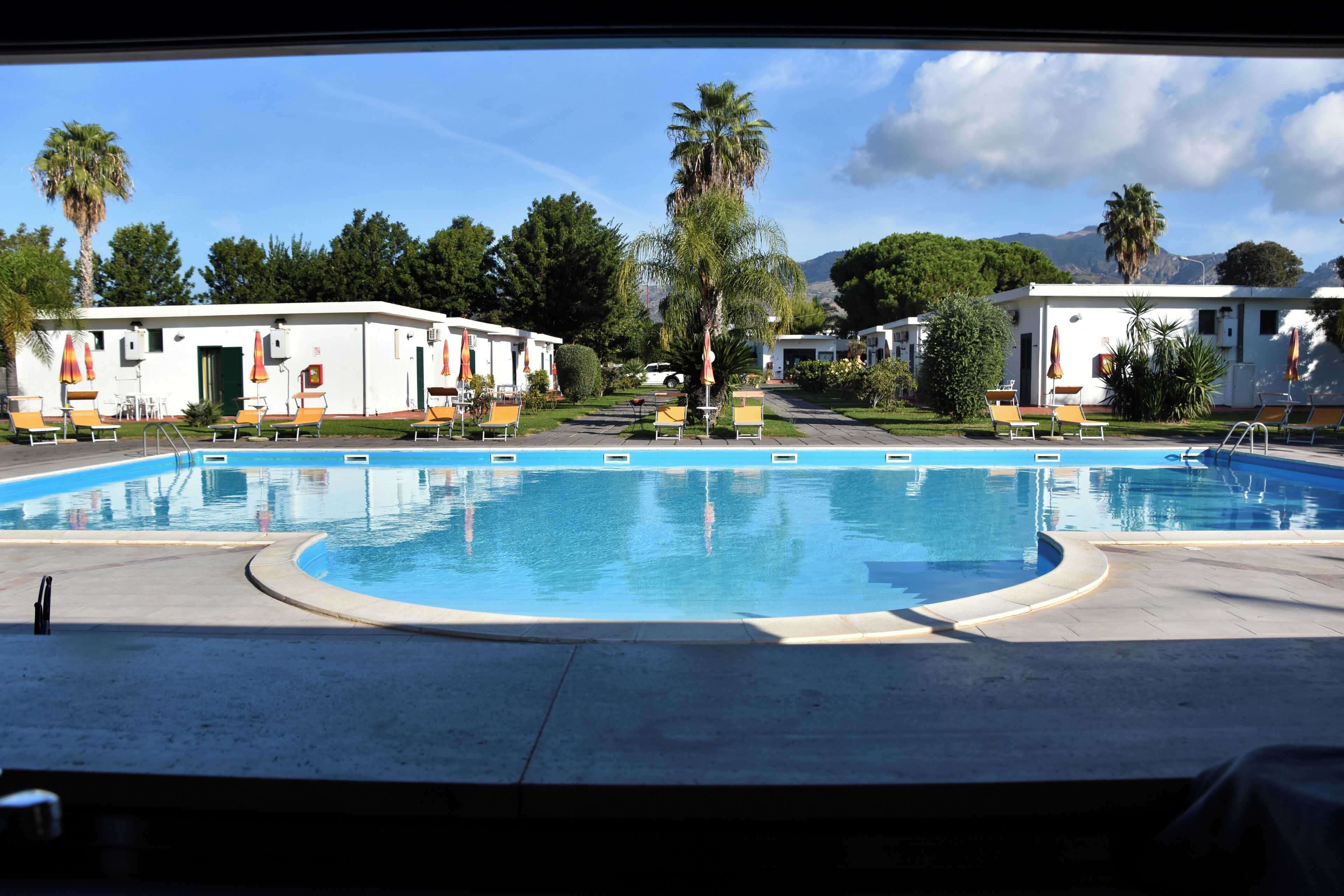 Une piscine entourée de bâtiments et d’arbres, avec des chaises longues et des parasols.