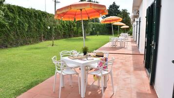 An outdoor patio with white tables and chairs, orange umbrellas, and a green hedge in the background.
