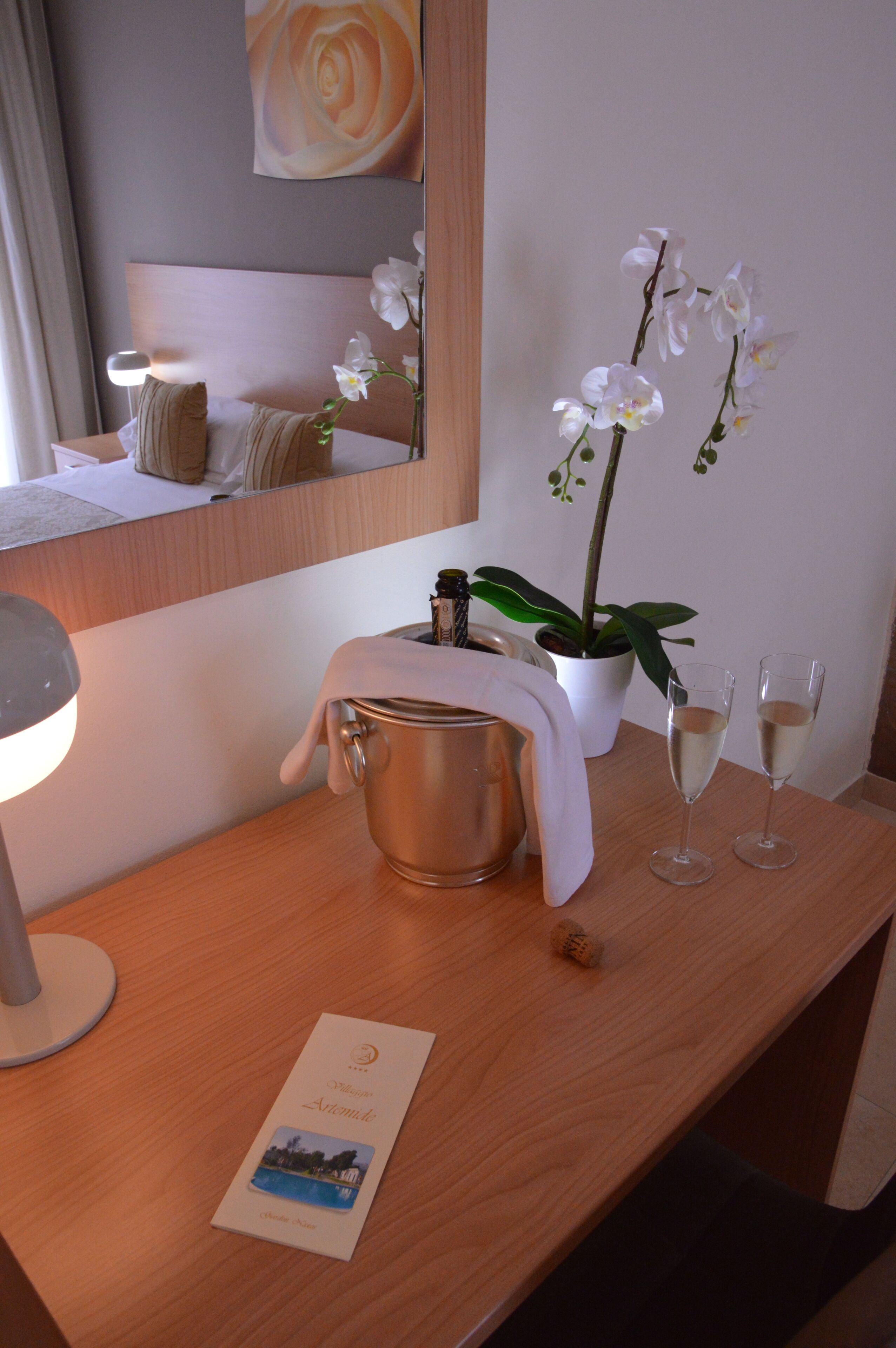 A hotel room with a wooden desk, a metal bucket, two glasses of white beverage, a white orchid, and a framed picture.