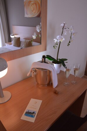 Habitación de hotel con un escritorio de madera, un cubo metálico, dos vasos de una bebida blanca, una orquídea blanca y un cuadro enmarcado.