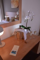 Habitación de hotel con un escritorio de madera, un cubo metálico, dos vasos de una bebida blanca, una orquídea blanca y un cuadro enmarcado.