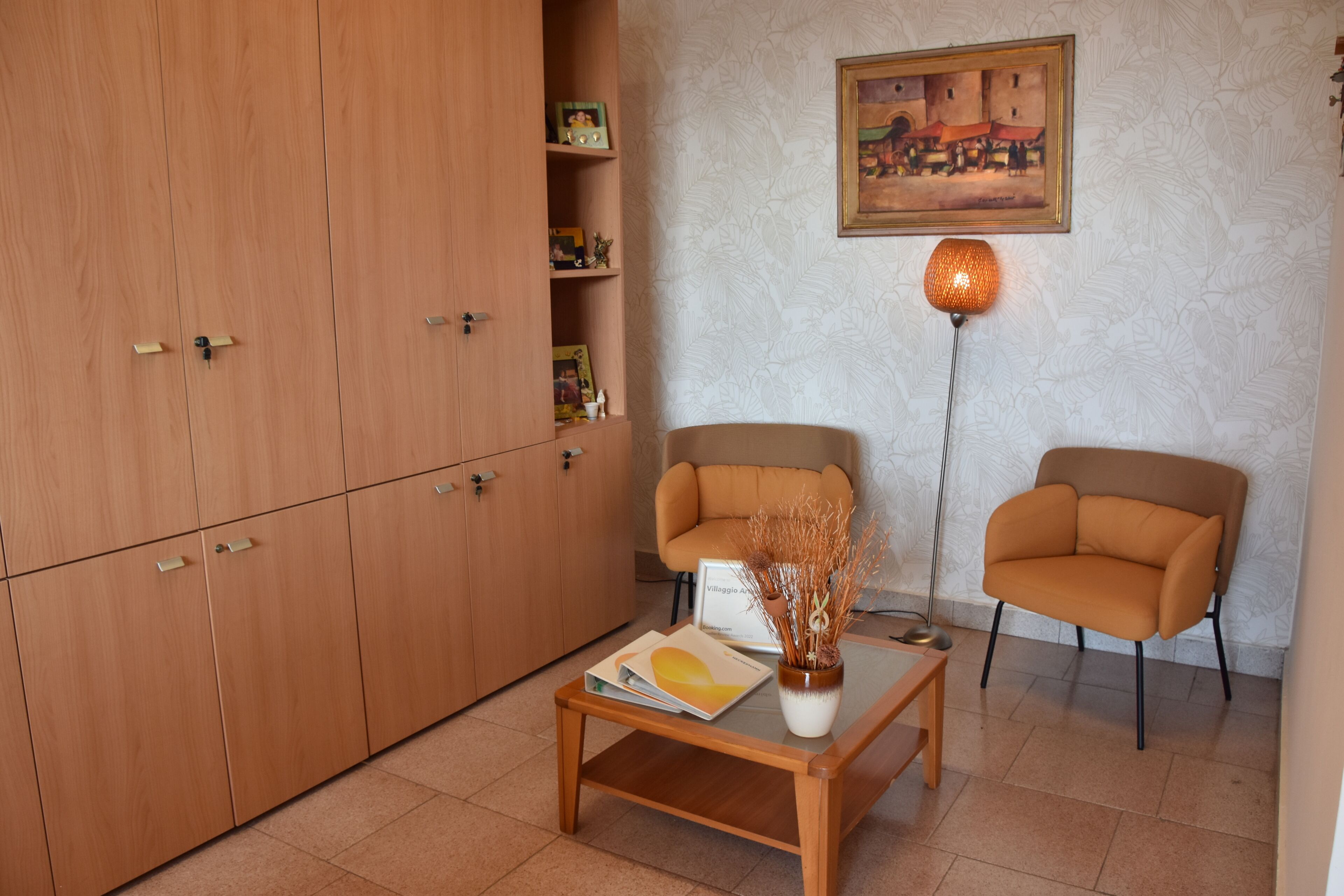 Una habitación con un armario de madera, dos sillones, una mesa de centro con una revista, un jarrón con plantas secas, una lámpara de pie y un cuadro en la pared.