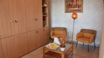Una habitación con un armario de madera, dos sillones, una mesa de centro con una revista, un jarrón con plantas secas, una lámpara de pie y un cuadro en la pared.