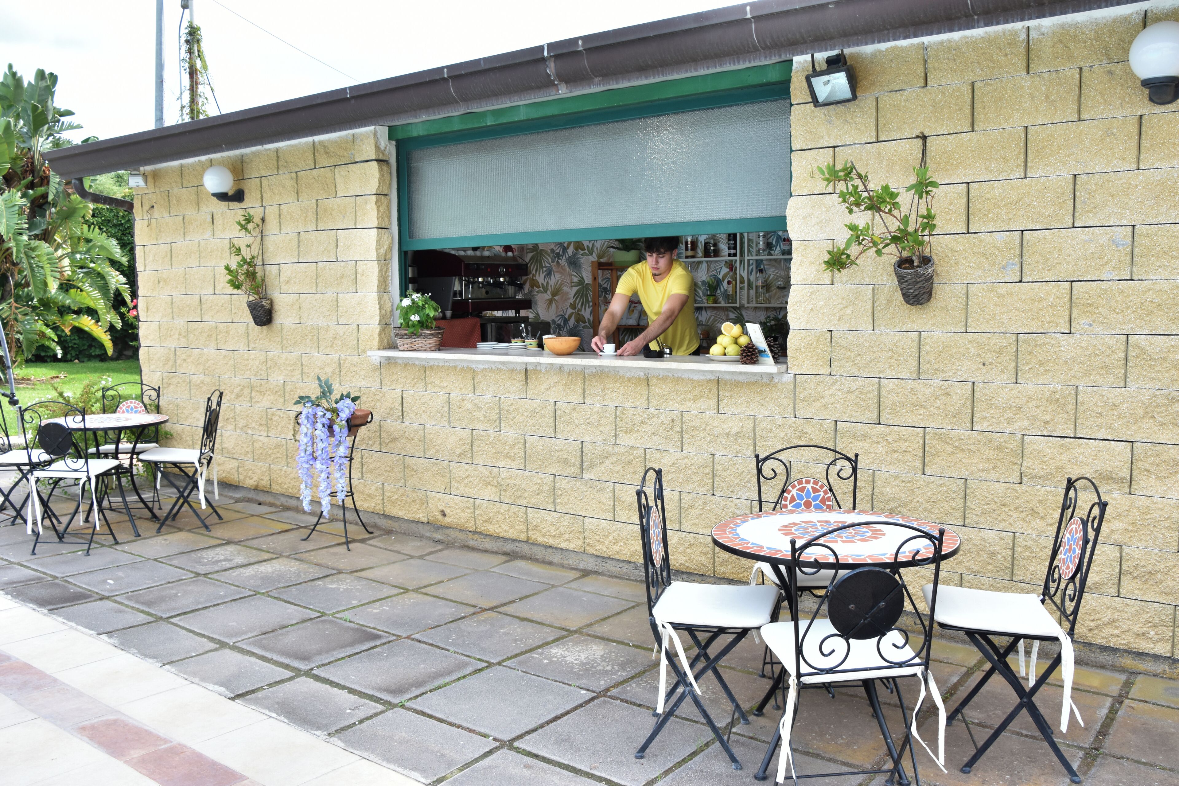 An outdoor patio with a brick wall, a window with a person working, and outdoor seating with metal chairs and tables.