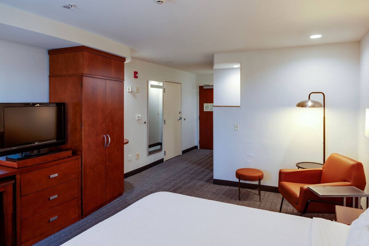 A hotel room with a bed, a television, a wooden wardrobe, a desk, an orange chair, and a lamp.