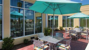 An outdoor patio with tables and chairs, large umbrellas, and a building with large windows in the background. - Hilton Garden Inn Houston-Pearland (Pearland)