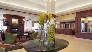 A lobby with a round glass table, green chairs, a fireplace, and a reception desk. - Hilton Garden Inn Houston-Pearland (Pearland)
