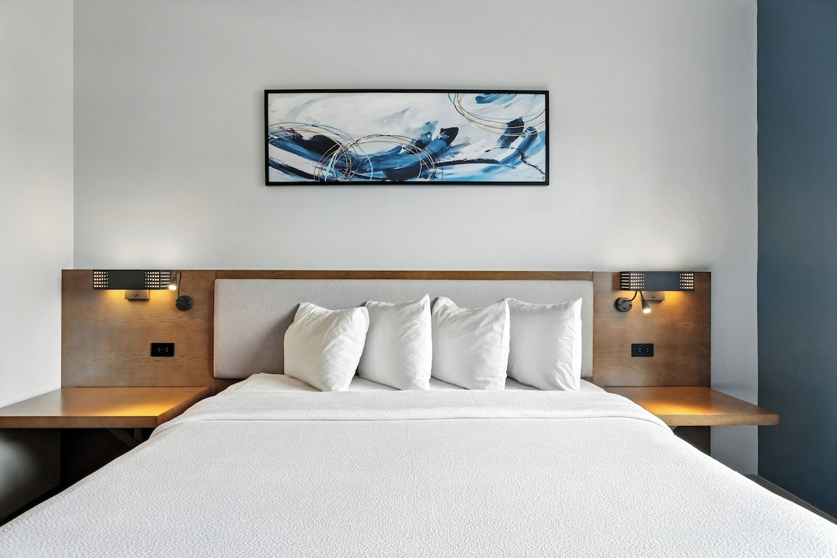 Ein modernes Hotelzimmer mit einem großen Bett, zwei Nachttischen mit Lampen, einer Kopfteil und einem Wandbild mit abstrakter Kunst.