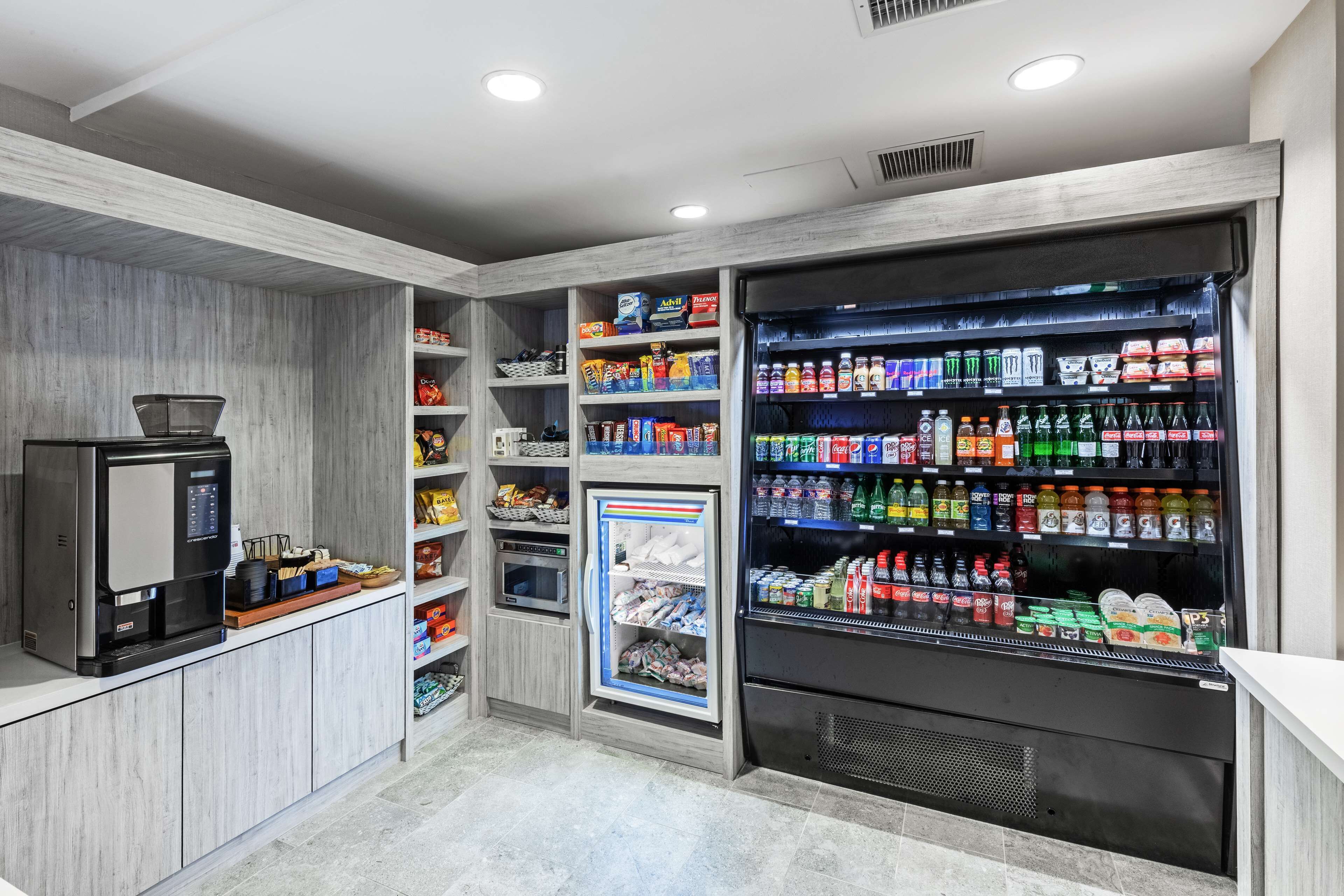 Zona de quioscos de autoservicio con máquina de café, dispensador de bebidas refrigeradas y estantes llenos de aperitivos y bebidas.