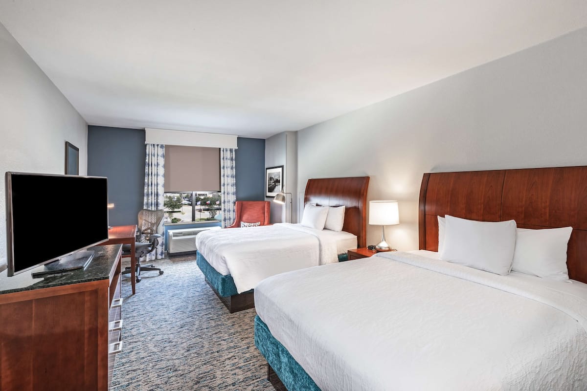 Ein Hotelzimmer mit zwei Betten, einem Fernseher, einem Schreibtisch und einem Fenster mit Vorhängen.
