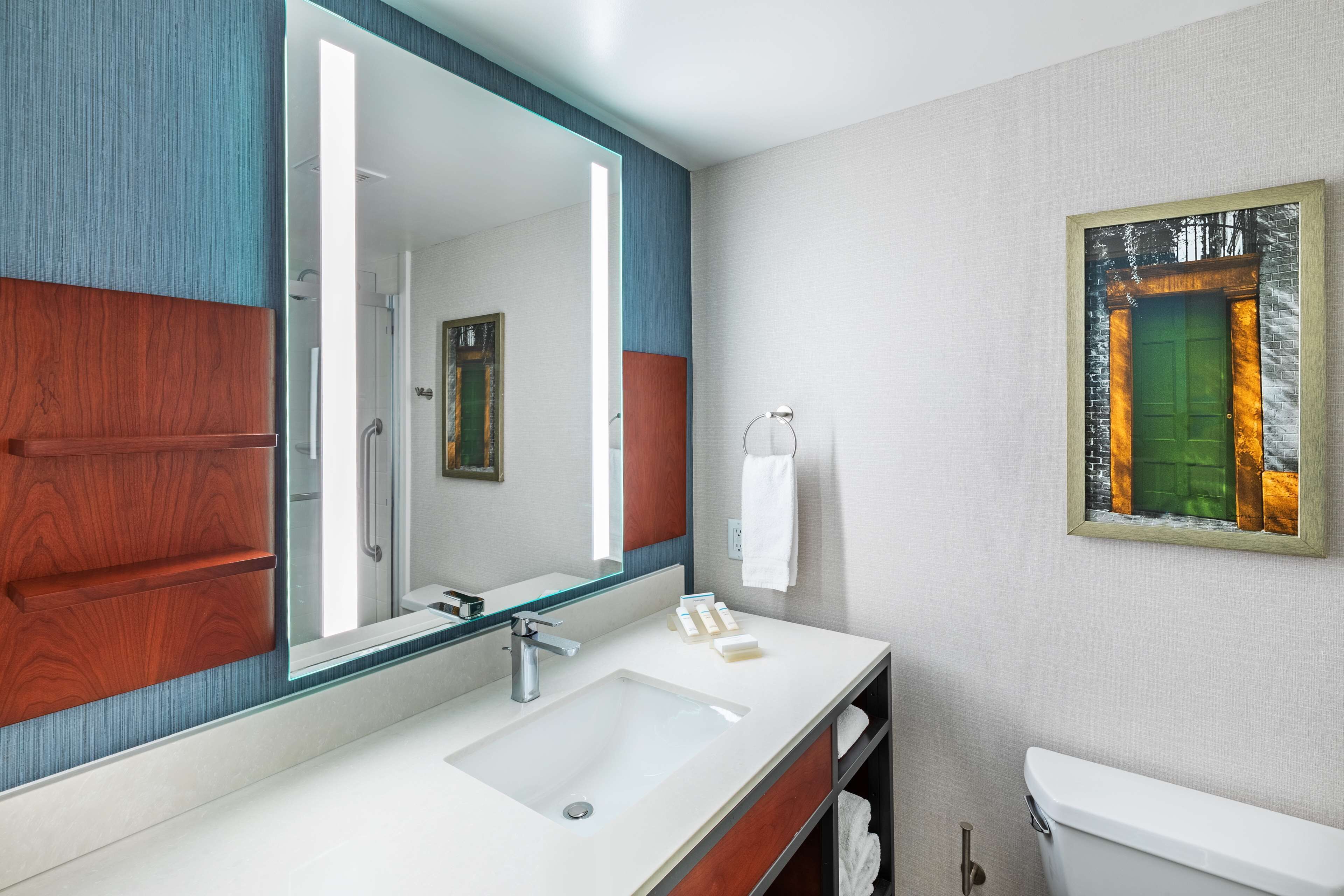 A modern bathroom with a large mirror, white sink, and a framed picture on the wall.