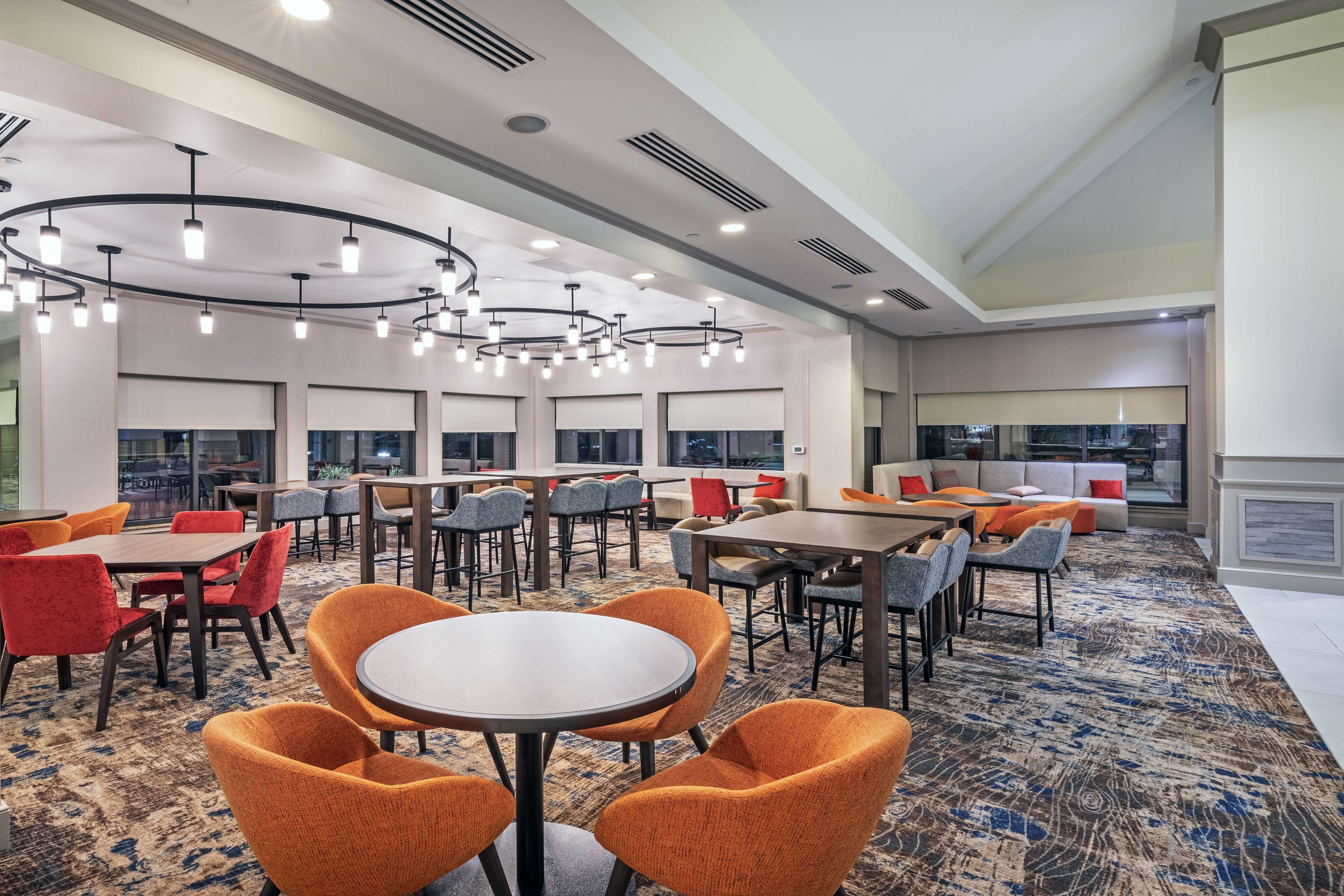 Un hall d’hôtel moderne avec des tables rondes, des chaises orange et un tapis à motifs.