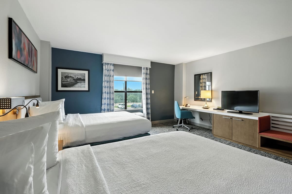 Ein Hotelzimmer mit zwei Betten, einem Schreibtisch, einem Fernseher und einem fensterbepflanzten Fenster.