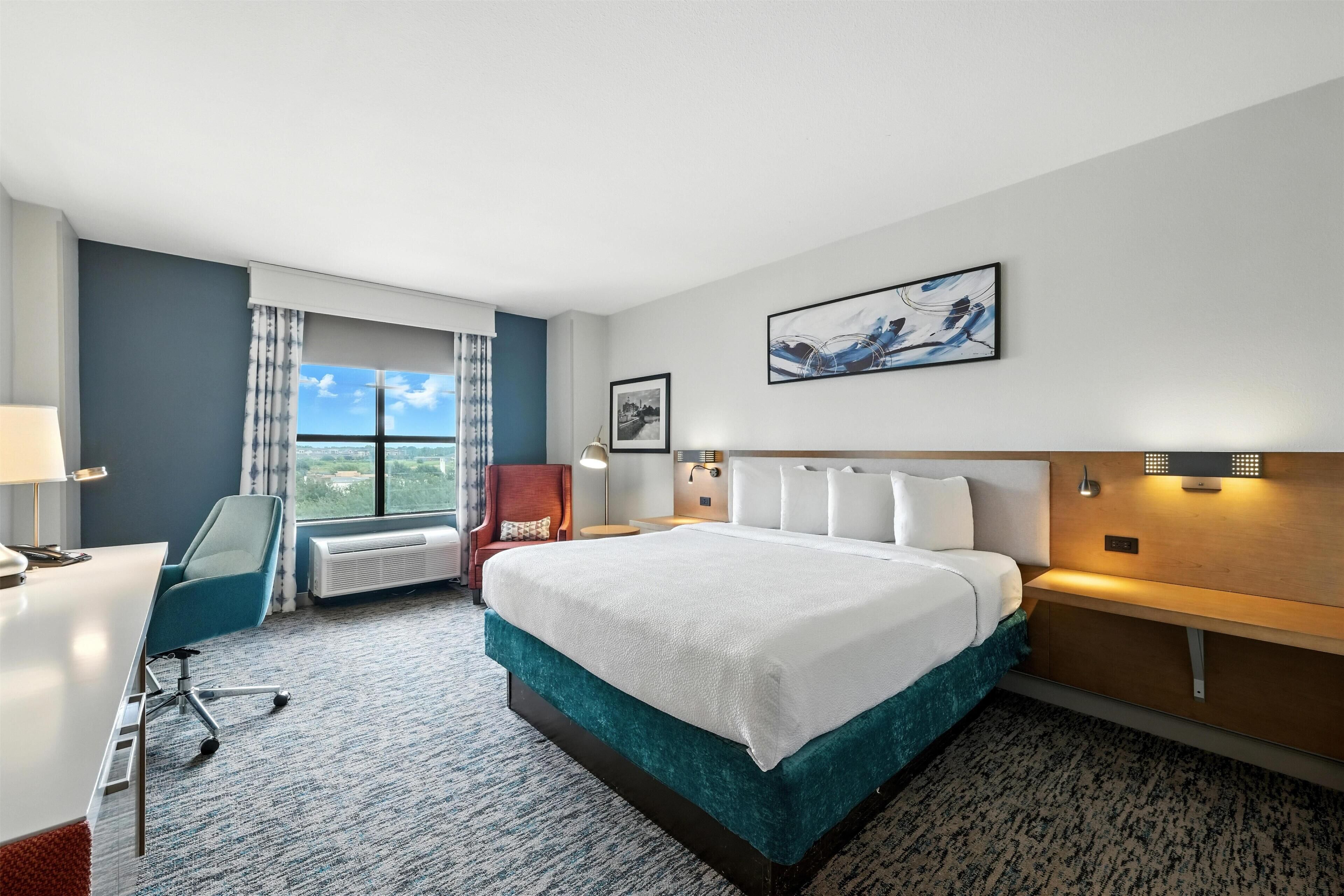 A hotel room with a bed, desk, chair, and a large window with a view.