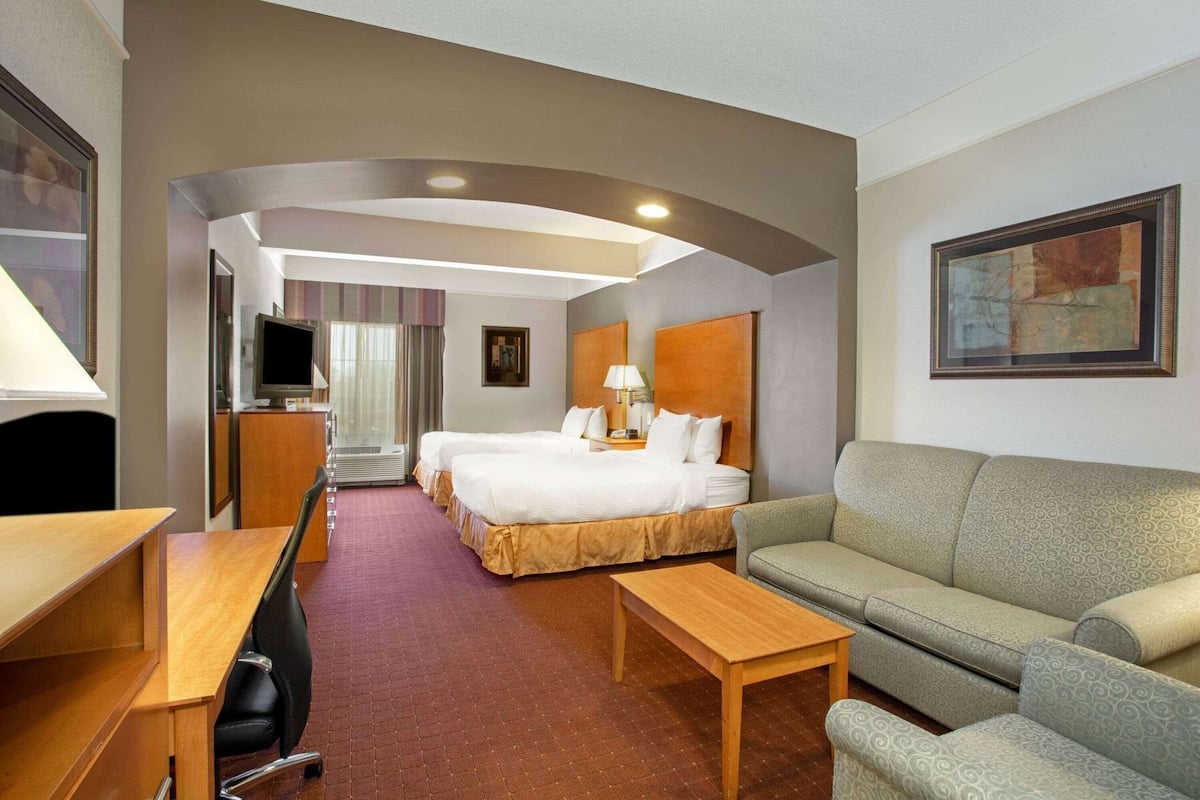 A hotel room with a bed, a sofa, a desk, a TV, and a painting on the wall.