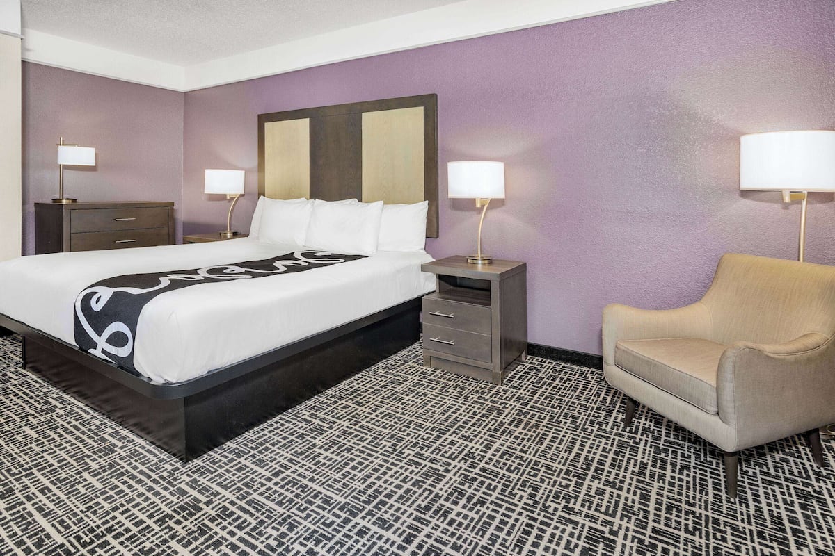 A hotel room with a large bed, a bedside table with a lamp, a chair, and a patterned carpet.