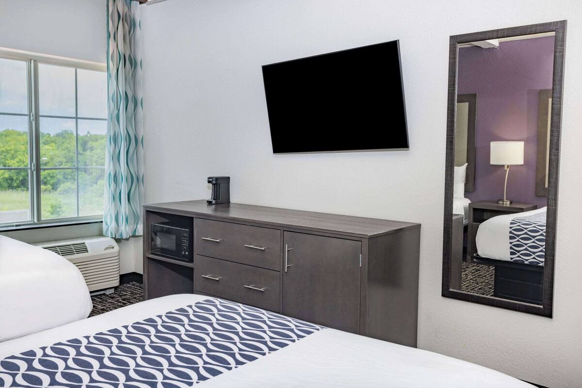 A hotel room with a bed, a flat-screen TV, a minibar, and a large window with curtains.