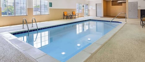 An indoor swimming pool with clear blue water, a tiled floor, and a ceiling painted in teal. There are two windows on the wall, a door, and a small seating area with chairs.