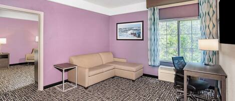 A hotel room with a purple wall, a beige sofa, a small desk with a chair, a window with curtains, and a framed picture on the wall.