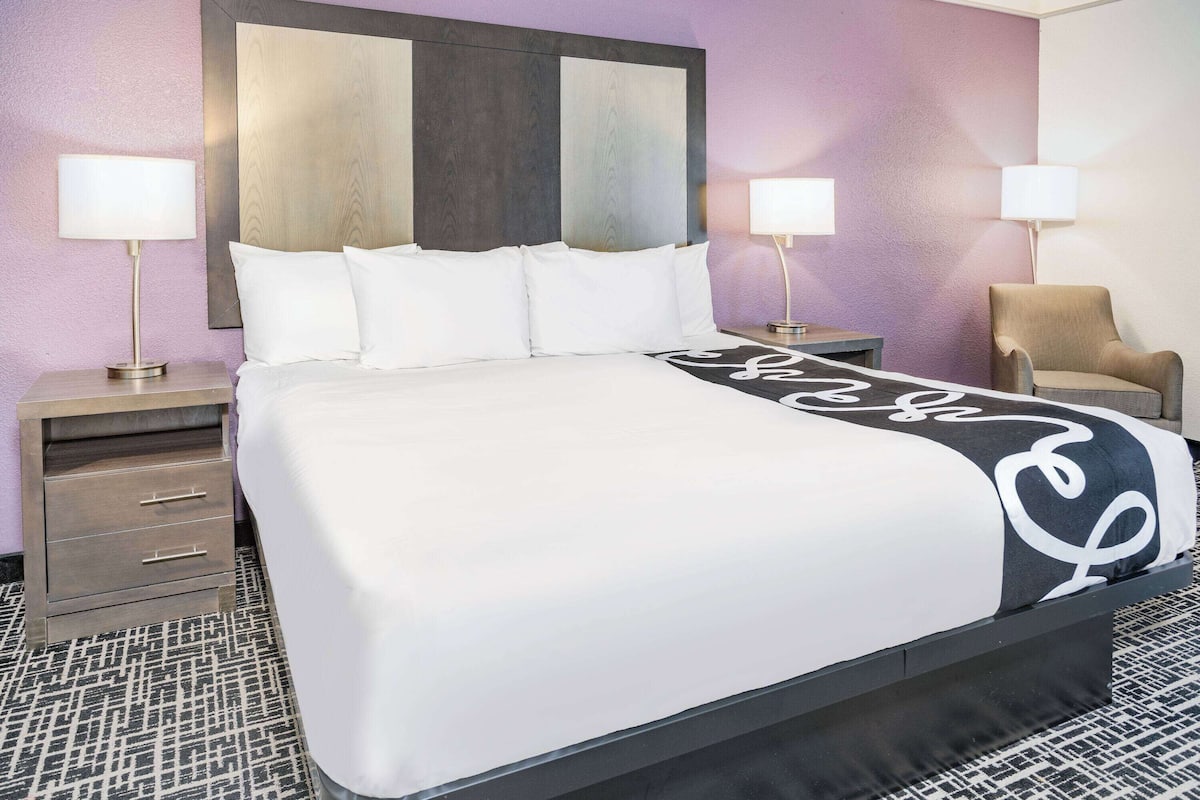 A hotel room with a large bed, two bedside tables, a chair, and a headboard with a wooden panel.