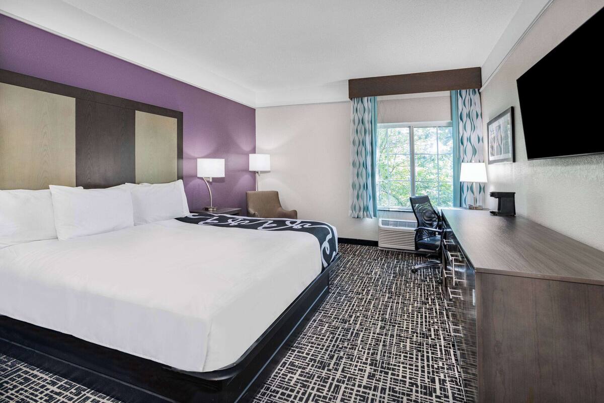 A hotel room with a large bed, a desk, a chair, a TV, and a window with curtains.