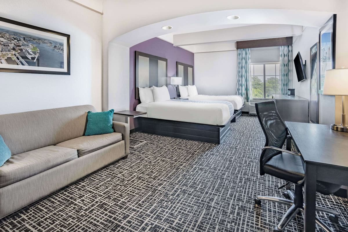 A hotel room with a bed, a desk, a chair, a sofa, and a large window with curtains.
