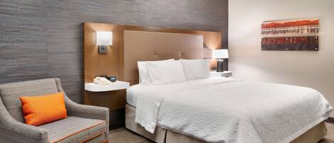 Habitación de hotel con cama, una silla, mesita de noche, lámpara y un cuadro en la pared.