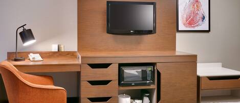 A hotel room with a wooden desk, a flat-screen TV, a microwave, a coffee maker, and a framed abstract painting on the wall.