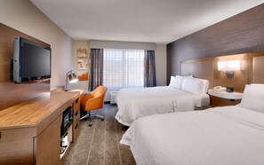 A hotel room with two beds, a desk with a flat-screen TV, a chair, a bedside table with a lamp, and a window with curtains.