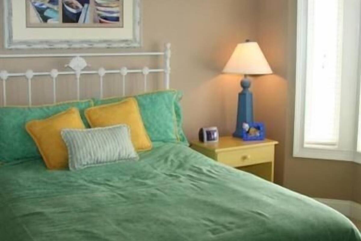 A bed with green bedding and yellow pillows, a bedside lamp, a small bedside table with a clock, and a framed picture above the bed.