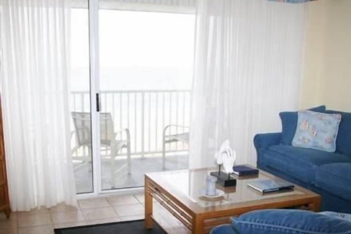 A living room with a glass door leading to a balcony, a blue sofa, a wooden coffee table, and a striped armchair.