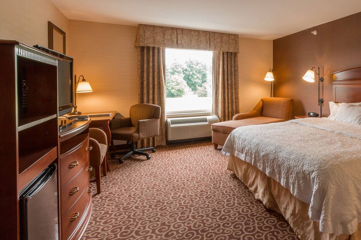 Habitación de hotel con una cama grande, un escritorio con silla, un aparador y una ventana con cortinas.