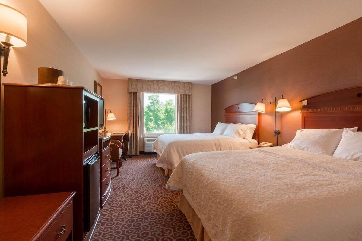 Una habitación de hotel con dos camas, un escritorio con silla, cómoda y ventana con cortinas.