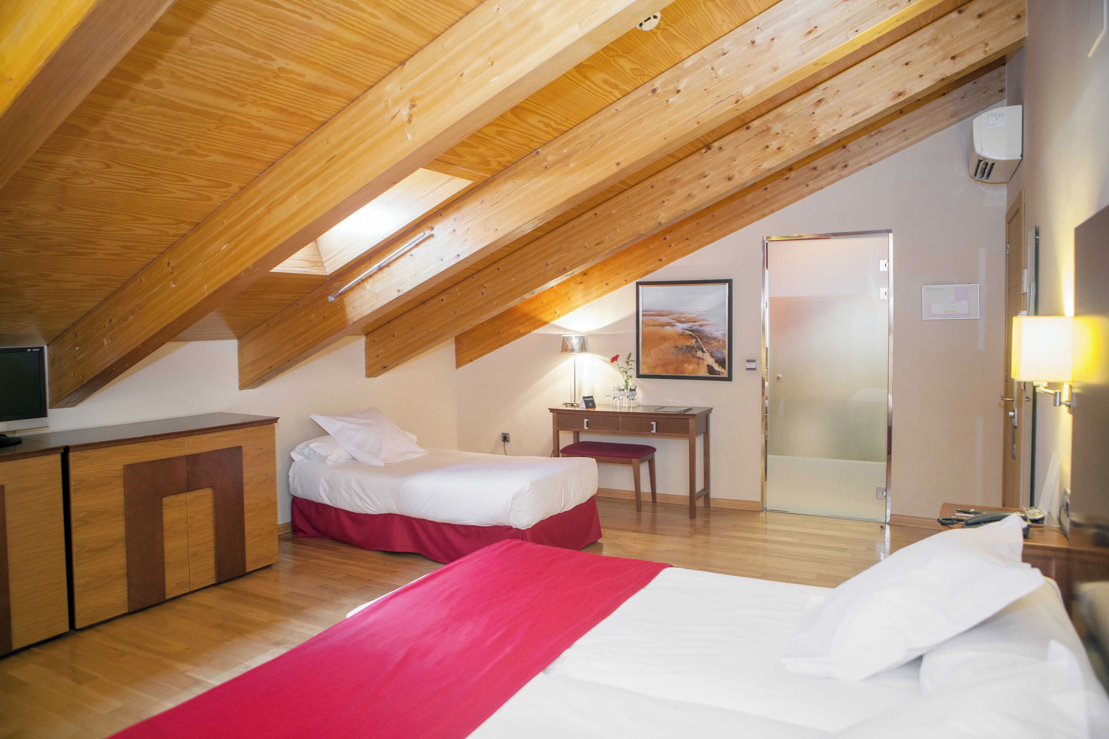 Una habitación de hotel con techo inclinado, vigas de madera, una cama con colcha roja y blanca, un escritorio con lámpara y una puerta que da a otra habitación.