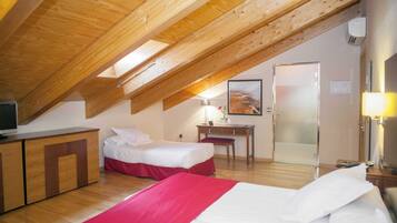 A hotel room with a sloped ceiling, wooden beams, a bed with a red and white bedspread, a desk with a lamp, and a door leading to another room.