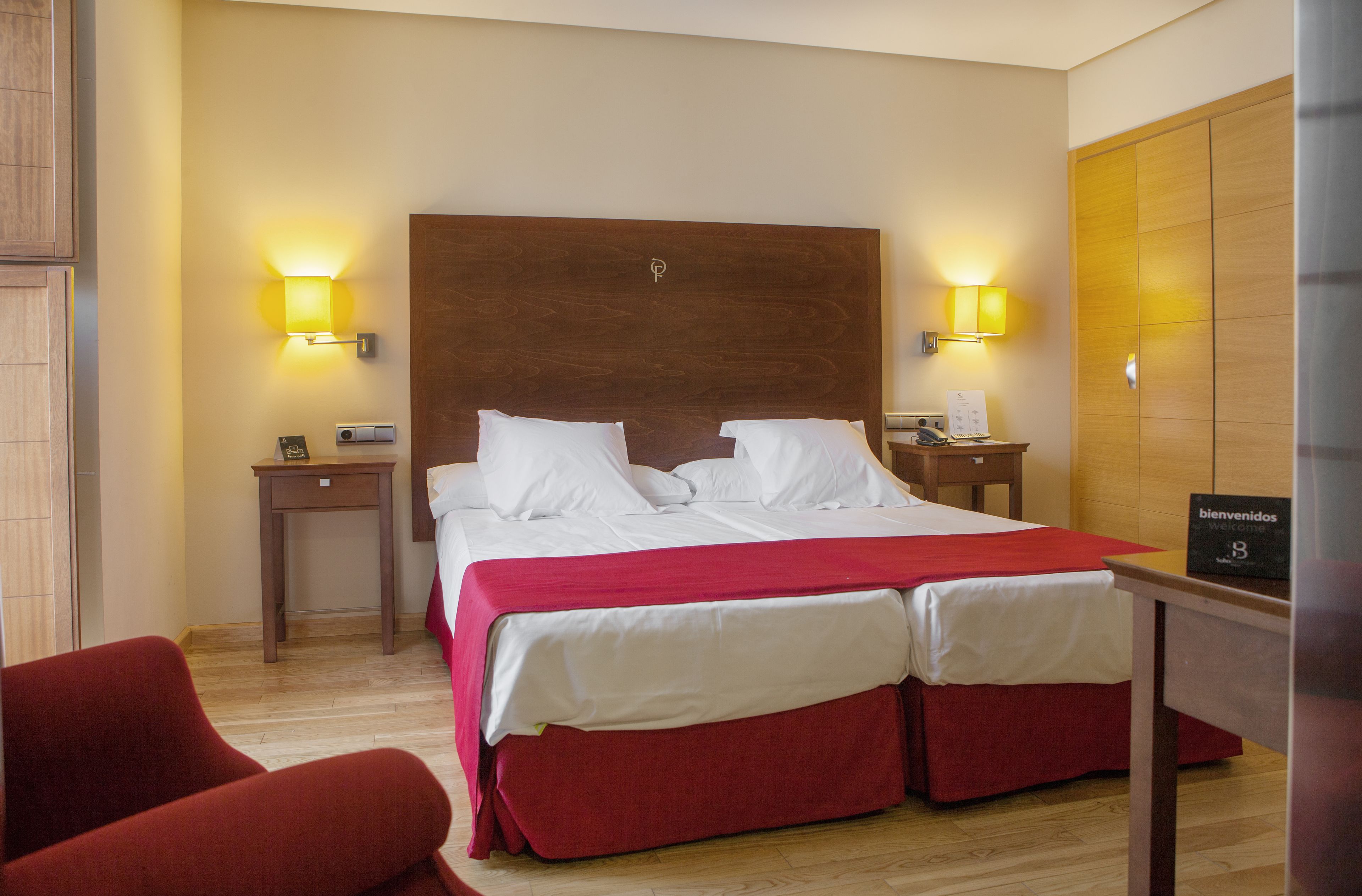 A hotel room with a wooden headboard, two beds with white linens, red bedspreads, two bedside tables with lamps, a red chair, and a 'bienvenidos welcome' sign.