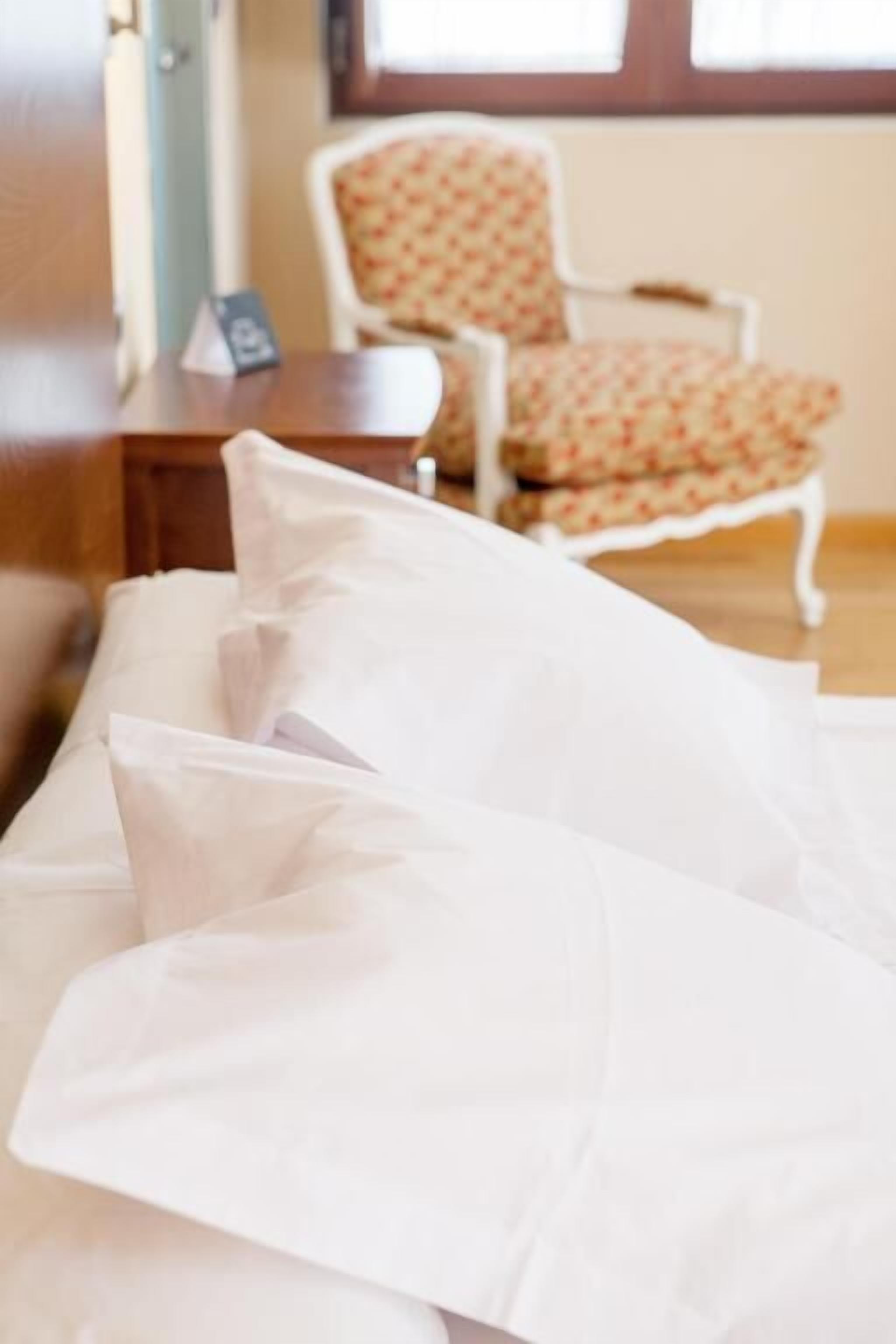 Una cama bien tendida con sábanas blancas en una habitación con una silla estampada con motivos florales y una mesita de noche con una lámpara.