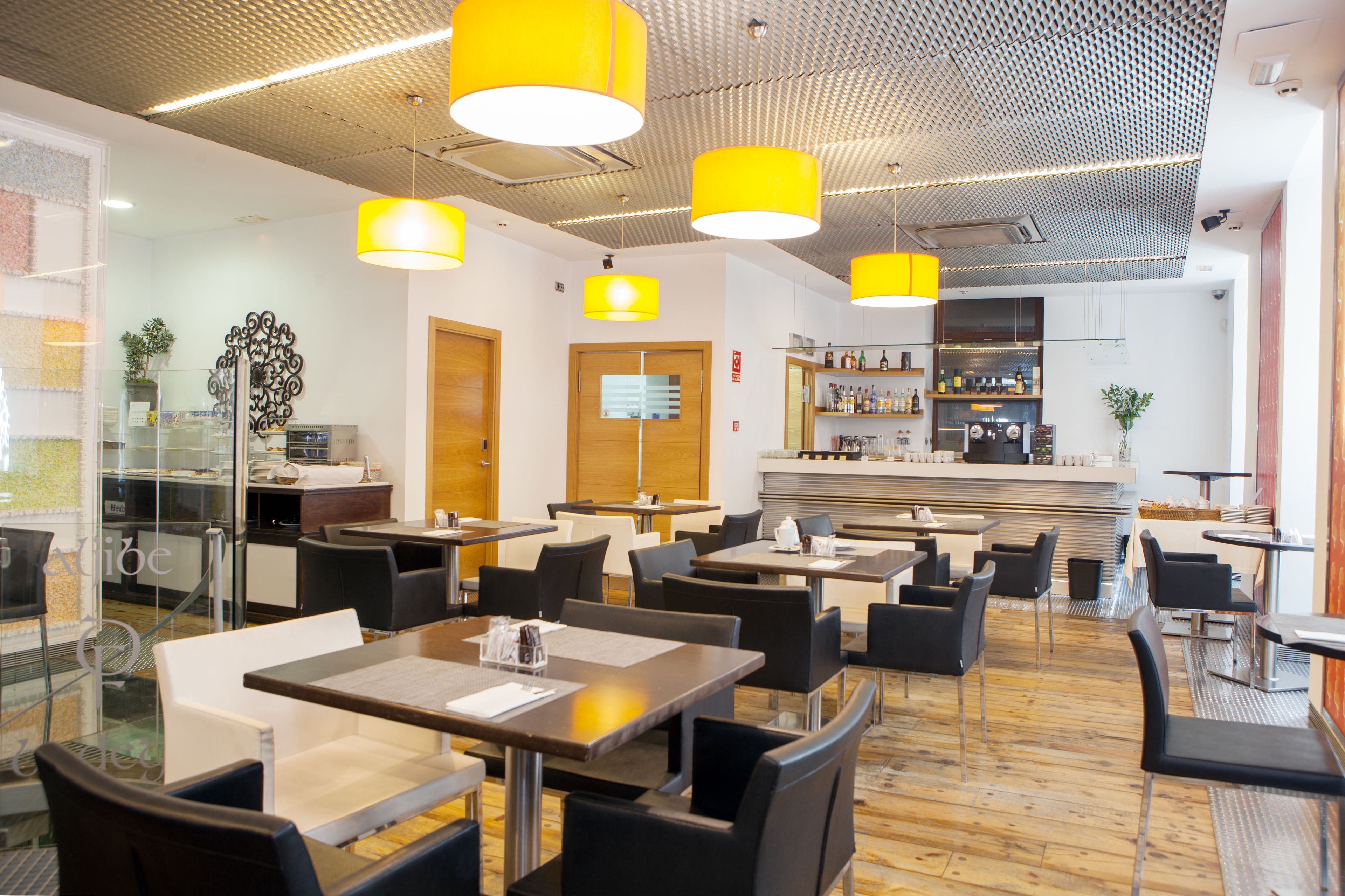 Ein modernes Restaurant mit mehreren Tischen und Stühlen, gelben Pendelleuchten und einer Bar im Hintergrund.