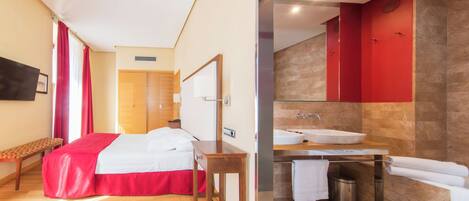 A hotel room with a bed, a bedside table, a bench, a mirror, and a bathroom with a sink and a bathtub.