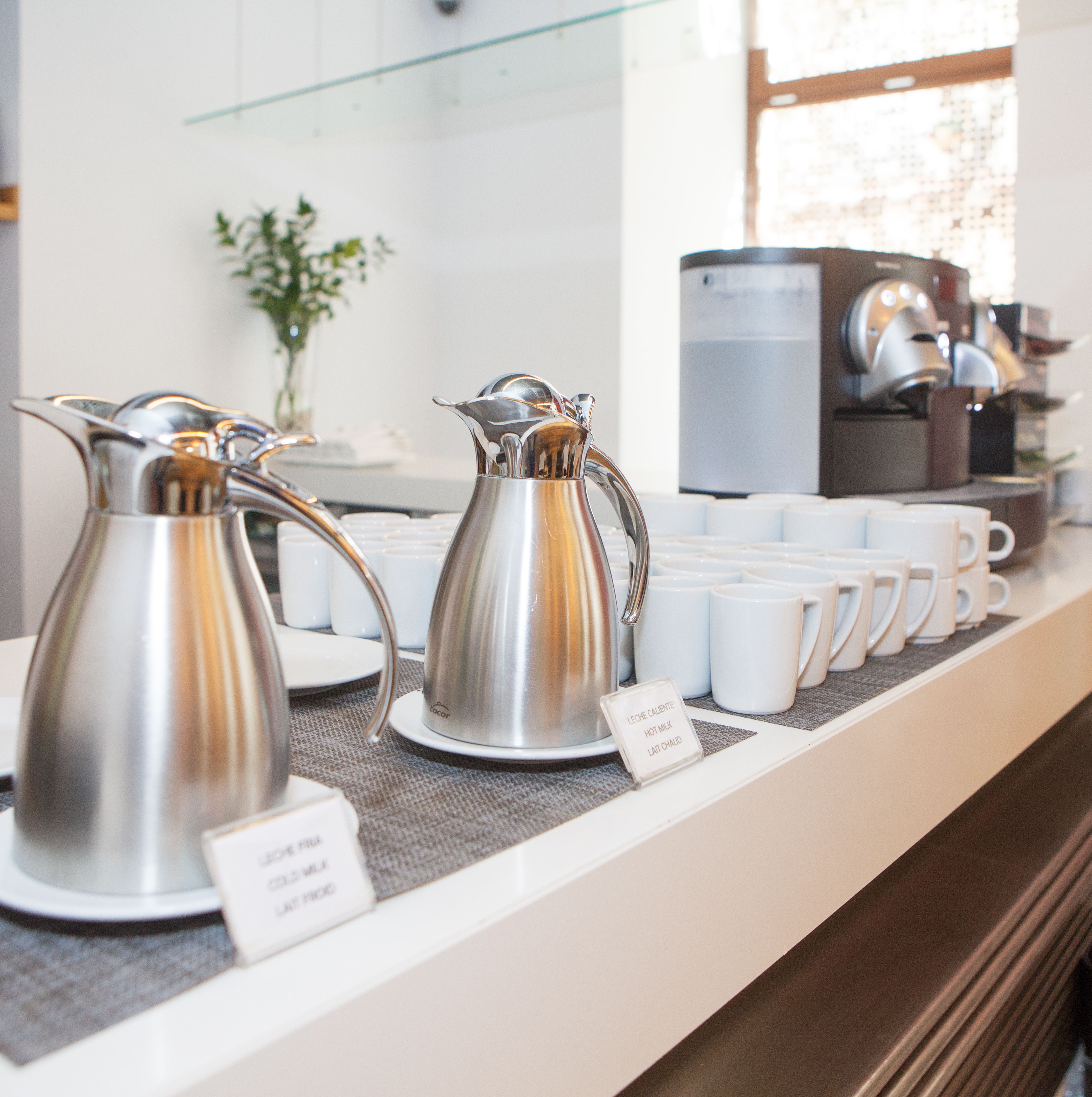 A coffee station with two silver coffee pots and several white coffee cups on a counter.