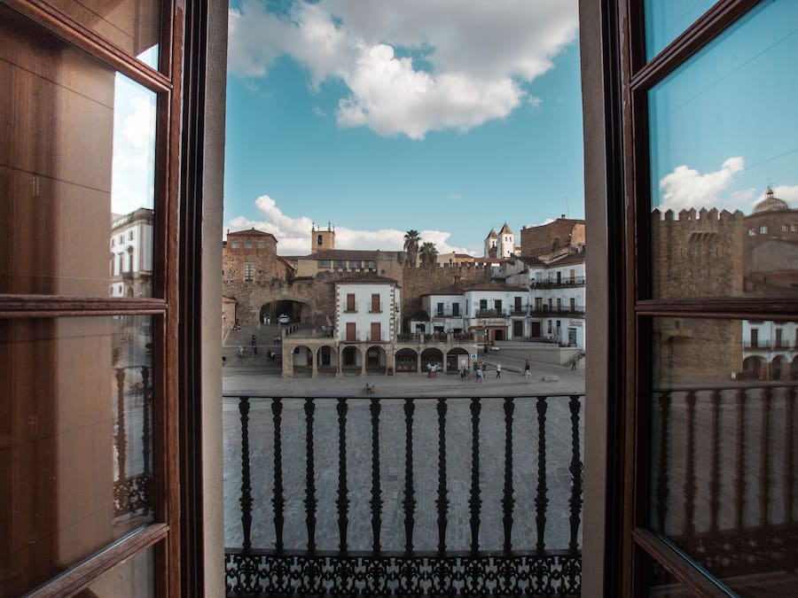 Vista desde una ventana hacia una plaza histórica con edificios, una fuente y cielo despejado.