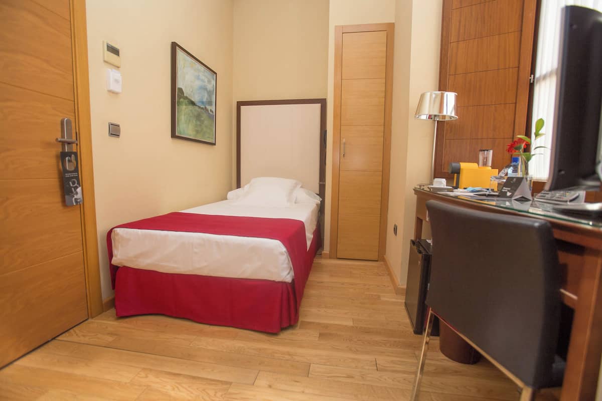 A hotel room with a bed, a desk, a chair, a TV, a lamp, and a door.
