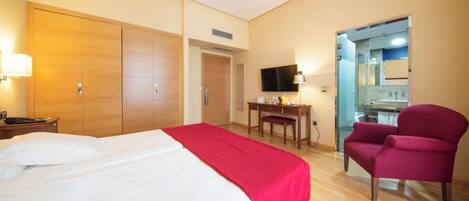 A hotel room with a bed, a desk, a chair, a TV, and a bathroom visible through a glass door.
